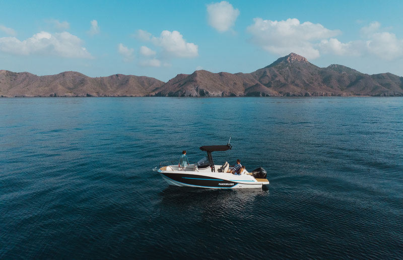 Bateau QuickSilver en mer avec deux personnes à bord, sous un ciel bleu et face à des montagnes à l’horizon.